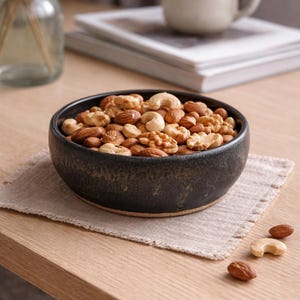 May include: A dark ceramic bowl filled with a mix of nuts, including almonds, cashews, and walnuts. The bowl sits on a beige linen napkin on a wooden table. A white mug and books are in the background.