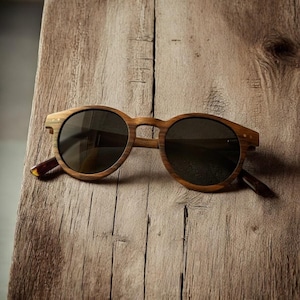 May include: Round-framed sunglasses with a natural wood grain finish. The sunglasses have dark lenses and a classic design. The arms of the sunglasses are a darker brown color. The sunglasses are resting on a wooden surface.