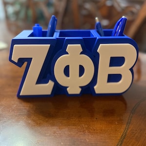 May include: A blue desk organizer with white lettering spelling out "ZΦB". The organizer has multiple compartments holding blue pens. The design is a bold, three-dimensional style, suitable for a desk or office setting.