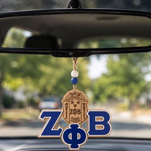 May include: A wooden car hanging ornament featuring the Greek letters ZPhiB in blue and white, suspended by a jute twine string with one white and one blue bead. The design includes a detailed crest with the text ZPhiB and the year 1920 engraved.