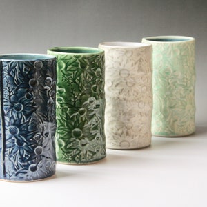 May include: Four ceramic vases in a row, each with a different colour and floral pattern. The colours are blue, green, off-white, and light green. The vases are cylindrical and have a textured surface.
