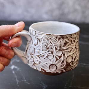 Może przedstawiać: Biały kubek ceramiczny z wypukłym wzorem kwiatowym. Kubek ma brązową glazurę i prezentuje szczegółowy wzór białych kwiatów i liści.