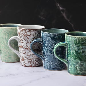 May include: Four ceramic mugs arranged in a row, each featuring a distinct embossed floral pattern. The mugs are coloured in light green, white with brown accents, dark blue, and a vibrant green, all with a glossy finish and curved handles.
