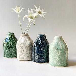 May include: Four ceramic vases in green, cream, and blue, each with a unique embossed floral design. One vase holds white flowers. The vases are arranged in a row, showcasing their varying textures and colours.