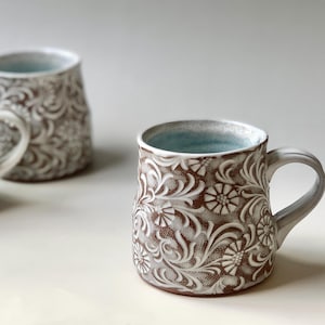 May include: Three ceramic mugs with a floral pattern. The mugs are brown with white embossed floral designs. The interior of the mugs is a light blue colour. The mugs have a handle.