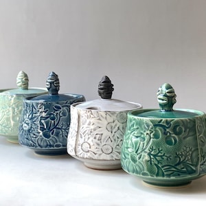May include: Four ceramic lidded jars with unique floral designs. The jars are in shades of blue, green, and white. Each jar has a different knob on the lid.