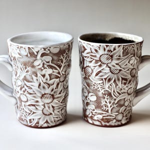 Może przedstawiać: Dwa ceramiczne kubki z kwiatowym wzorem. Kubki są brązowe z białymi kwiatowymi wzorami i białymi uchwytami. Jeden kubek jest wypełniony ciemnym płynem. Kubki stoją na białej powierzchni.