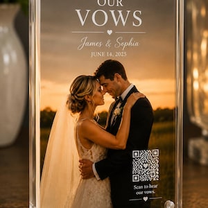 May include: Clear acrylic photo block with a wedding photo of a couple embracing at sunset. Text reads "OUR VOWS", "James & Sophia", and "JUNE 14, 2025". A QR code is also visible. The block is held together with silver screws.