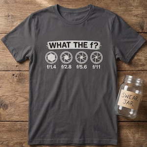 Könnte beinhalten: Dunkelgraues T-Shirt mit dem Text "WHAT THE f?" über Kamera-Blenden-Symbolen und Blendenzahlen. Rechts befindet sich ein Glas mit der Aufschrift "SWEAR JAR". Das T-Shirt liegt auf einer Holzoberfläche.