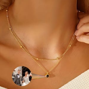 May include: A gold-colored, layered necklace with a small, round, black pendant. The necklace has a delicate chain design with small, round beads. A small, circular image of a couple is projected onto the necklace.