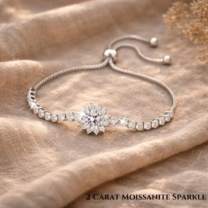May include: A silver bracelet with a central flower-shaped design, surrounded by smaller, sparkling stones. The bracelet has an adjustable clasp. The text at the bottom reads "2 CARAT MOISSANITE SPARKLE".