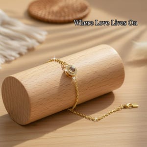 May include: A gold-colored bracelet with a circular pendant, displayed on a wooden cylinder. The pendant has a central stone surrounded by smaller stones. The text "Where Love Lives On" is visible.