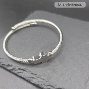 May include: A silver-toned cable wire bracelet featuring a central cross charm flanked by two small metallic beads. The bracelet has an open-ended design with ribbed end caps. The text FAITH-INSPIRED is visible in the upper right corner.