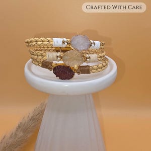 May include: Three braided leather bracelets stacked together, each featuring a central oval druzy stone in white, yellow, and dark brown. The stones are set in gold-tone metal, with matching thread accents. The text CRAFTED WITH CARE is visible.