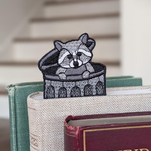 Pode incluir: Marcador de livros bordado de guaxinim num design de caixote do lixo. O marcador de livros apresenta um guaxinim cinzento e branco a espreitar de um caixote do lixo preto e cinzento. O marcador de livros é colocado entre as páginas de uma pilha de livros.
