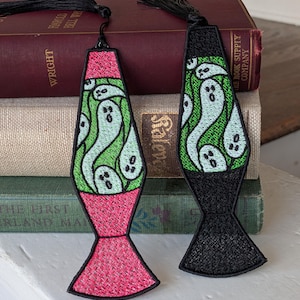 Könnte beinhalten: Zwei gestickte Lesezeichen in Form von Lavalampe, eines mit rosa Basis, das andere schwarz. Jedes hat ein grünes Innere mit weißen Geistermotiven und eine schwarze Quaste. Sie liegen auf einem Stapel alter Bücher.