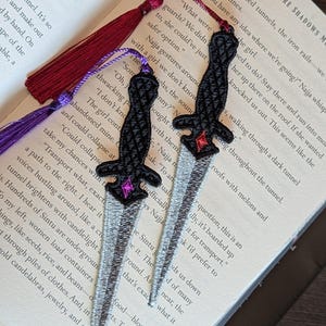 Puede incluir: Dos marcapáginas bordados con forma de daga, con mangos negros, hojas plateadas y borlas decorativas en burdeos y morado. Los marcapáginas están colocados sobre un libro abierto con texto impreso.