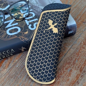Puede incluir: Estuche para gafas negro y dorado con un diseño de escamas de dragón y un emblema de dragón dorado. El estuche está hecho de un material texturizado y está diseñado para proteger las gafas. Unas gafas negras descansan sobre un libro.