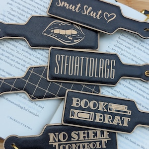 May include: A collection of black leather paddles with various phrases and designs etched in a lighter color. The phrases include "Smut Slut," "STFUATTOLAGG," "Book Brat," and "No Shelf Control." Some paddles have additional graphic elements.