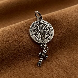 May include: A silver pendant with a circular design featuring the text "CHROME HEARTS" around the edge. The pendant has a cross charm hanging below. The design includes figures and a central sword.
