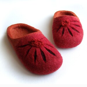 May include: A pair of red felt slippers with a flower design. The slippers have a brown felt lining and a small pom-pom in the center of the flower.
