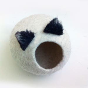 May include: A white felt cat bed with two black felt ears. The bed has a round opening for the cat to enter.