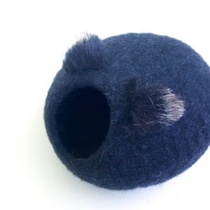 May include: A dark blue felt nesting ball with a small black and white fur pom-pom on top.