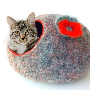K&ouml;nnte beinhalten: Ein graues und orangefarbenes Filzkatzenbett mit einem roten Blumendesign. Eine getigerte Katze sitzt im Bett.