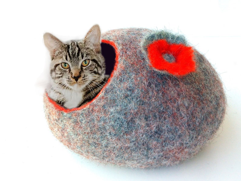 K&ouml;nnte beinhalten: Ein graues und orangefarbenes Filzkatzenbett mit einem roten Blumendesign. Eine getigerte Katze sitzt im Bett.
