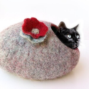 K&ouml;nnte beinhalten: Eine schwarze Katze schaut aus einem grauen und roten Filzkatzenbett mit einer roten Filzblume oben heraus.
