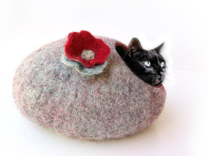 K&ouml;nnte beinhalten: Eine schwarze Katze schaut aus einem grauen und roten Filzkatzenbett mit einer roten Filzblume oben heraus.