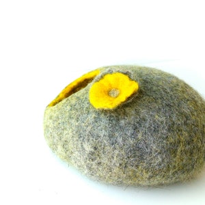 K&ouml;nnte beinhalten: Eine graue Filz-Nistkugel mit einer gelben Filzblume oben drauf. Die Kugel ist aus Wolle und hat eine weiche, flauschige Textur.