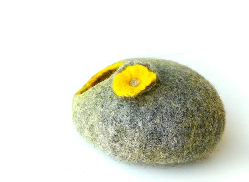 K&ouml;nnte beinhalten: Eine graue Filz-Nistkugel mit einer gelben Filzblume oben drauf. Die Kugel ist aus Wolle und hat eine weiche, flauschige Textur.