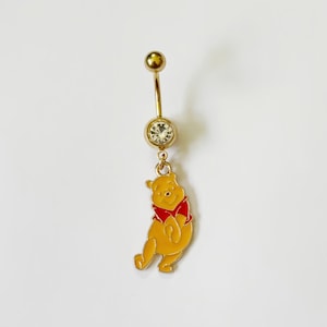 May include: Gold-colored belly button ring featuring a Winnie the Pooh charm. The charm is yellow with a red shirt. The top and bottom balls are gold, and the center has a clear gem.