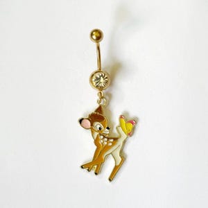 Puede incluir: Piercing para el ombligo dorado con un dije de Bambi. El dije es un ciervo de dibujos animados con detalles marrones, blancos y amarillos. Las bolas superior e inferior del anillo son doradas.