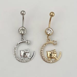 May include: Two belly button rings. One is silver-toned, the other gold-toned. Each features a curved crescent moon design with a small cat silhouette and a sparkling gem. The top and bottom gems are clear.