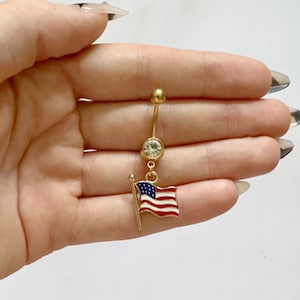 May include: Gold-colored belly button ring with a clear gem and a small American flag charm. The flag is red, white, and blue. The ring is designed for body piercing.