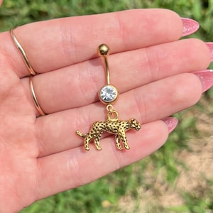 May include: Gold-colored belly button ring with a clear gem and a small, detailed leopard charm. The leopard has black spots and is in a walking pose. The ring has a curved barbell design.