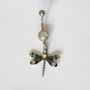 May include: A silver-toned belly button ring with a dragonfly charm. The dragonfly has a detailed design with colorful gemstones in shades of pink, blue, green, and clear. The top and bottom balls of the ring are also clear.