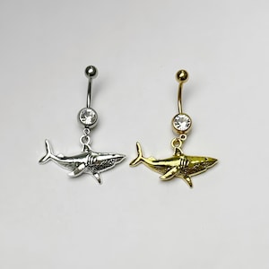 May include: Two belly button rings. One is silver-toned with a clear gem and a shark charm. The other is gold-toned with a clear gem and a shark charm. Both have a curved barbell design.