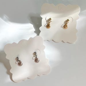 May include: Two pairs of seashell-shaped earrings. One pair is gold-coloured, and the other is silver-coloured. Each earring has a small ball at the top and a dangling seashell charm. The earrings are displayed on white, cloud-shaped earring cards.