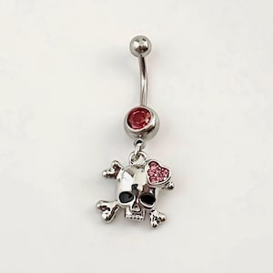 May include: A silver-colored belly button ring featuring a skull and crossbones charm with a pink heart and a red gem. The curved barbell has a small silver ball at the top and a larger red gem at the bottom.