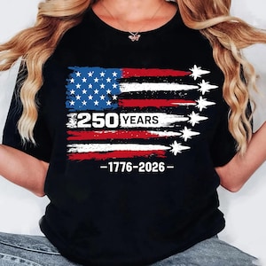 Könnte beinhalten: Ein schwarzes T-Shirt mit einer Grafik der US-Flagge im Used-Look und weißen Flugzeugsilhouetten, die Rauchfahnen hinterlassen. Der Text 250 YEARS und 1776-2026 ist in weißen Blockbuchstaben auf der Vorderseite des Kleidungsstücks gedruckt.
