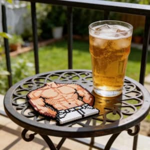 May include: A glass of beer with ice cubes sits on a decorative metal table next to a pixelated coaster shaped like a muscular torso. The coaster features shades of brown, white, and black. The table is outdoors on a sunny day.