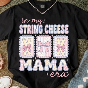 May include: Black t-shirt featuring the phrase "in my string cheese mama era" in pastel shades. The design incorporates string cheese and bow illustrations. A necklace is visible at the neckline.