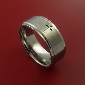 May include: A brushed silver wedding band with a cross design. The band is a simple, modern style.