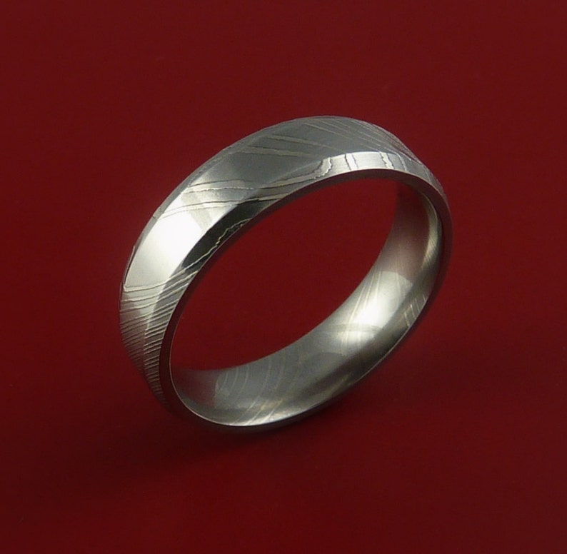 May include: A silver-toned wedding band with a unique, patterned design. The band is made of Damascus steel and has a polished finish.
