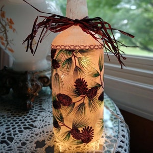 May include: A clear glass bottle with a brown ribbon and a pine cone and pine needle design. The bottle is lit from inside, creating a warm glow.