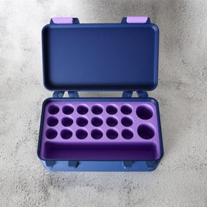 May include: An open, navy blue case with a purple interior. The case has a rectangular shape with rounded corners and a hinged lid. The interior features multiple circular compartments and a larger rectangular space.