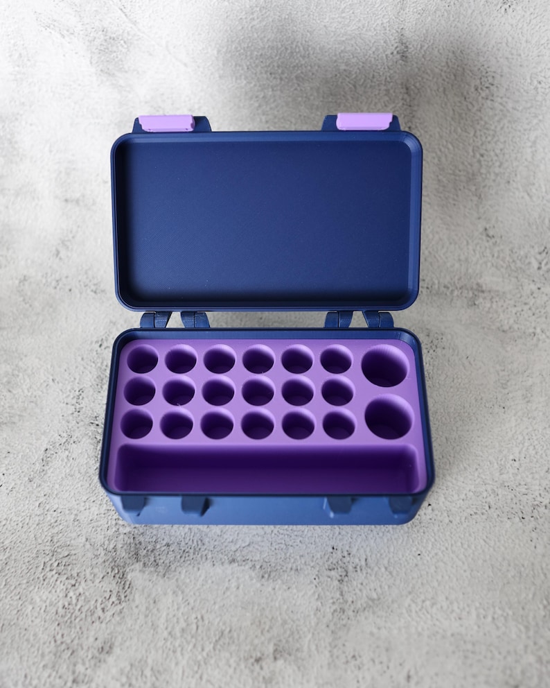 May include: An open, navy blue case with a purple interior. The case has a rectangular shape with rounded corners and a hinged lid. The interior features multiple circular compartments and a larger rectangular space.